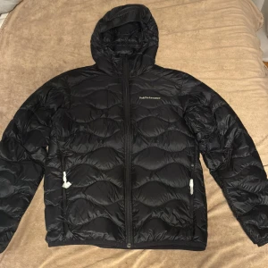 Svart dunjacka Peak Performance M - Helt ny Svart dunjacka från Peak Performance i storlek M. Jackan har huva, två sidofickor med dragkedja och snyggt quiltat mönster. Perfekt för kalla dagar med sin fluffiga och värmande design. Logotypen är broderad på bröstet och jackan stängs med dragkedja.