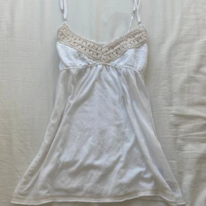 Vit kortklänning Abercrombie & Fitch - Supersöt vit kortklänning från Abercrombie & Fitch med justerbara smala axelband och broderad logga längst ner. Överdelen har beige virkade detaljer och smock baktill för extra komfort. Perfekt för varma dagar och chill vibes.