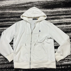 Vit hoodie från Polo Ralph Lauren - Vit hoodie från Polo Ralph Lauren med dragkedja, snörning i huvan och klassisk blå logga broderad på bröstet. Mjuk bomullsmix, kängurufickor och ribbade muddar. Perfekt för en clean och stilren look.