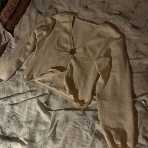 Beige croppad bolerotopp med knapp - Superfin beige bolerotopp med croppad passform och v-ringning. Toppen har en dekorativ guldig knapp framtill och långa ärmar med lätt puff. Materialet är mjukt och består av polyester och polyamid, vilket gör den skön att bära. Perfekt för dig som gillar stilrena och trendiga plagg.