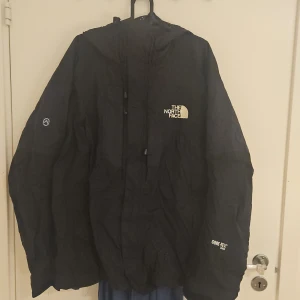 Svart vindjacka från The North Face - Svart vindjacka från The North Face Summit Series med huva och snörning. Jackan är tillverkad i Gore-Tex-material som står emot regn och vind. Logga på bröstet och ärmen, samt dragkedja framtill. Perfekt för dig som gillar outdoor och streetstyle.