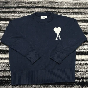 Mörkblå stickad tröja från Ami Paris - Mörkblå stickad tröja från Ami Paris med rund halsringning och ribbade muddar. På bröstet finns ett vitt hjärta med bokstaven A, vilket ger en unik och stilren look. Perfekt för dig som gillar klassiska men trendiga plagg.