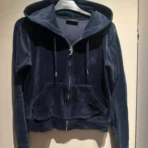Mörkblå  hoodie Juicy Couture - Mörkblå hoodie från Juicy Couture i mjuk velour med dragkedja framtill, huva och snörning. Klassisk känguruficka och diskret broderad logga på bröstet. Perfekt för en chill och trendig look.