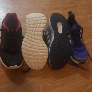 Adidas sneakers svart och blå - Två par Adidas sneakers, ett par i svart med röda detaljer och ett par i blått med vita ränder. Båda har snörning och platt sula, tillverkade i syntetmaterial med mesh-ytor för bra ventilation. Perfekta för dig som gillar sportig stil och vill ha bekväma skor.