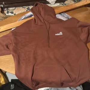 Brun Adidas hoodie med stor logga - Säljer en brun hoodie från Adidas med stor vit logga på ryggen och mindre logga på bröstet. Tröjan har huva med snörning och en stor magficka. Perfekt för dig som gillar streetwear och vill ha en skön och snygg look.