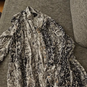 Mönstrad skjortklänning från KappAhl - Säljer en långärmad skjortklänning från KappAhl med coolt ormmönster i svart, grått och vitt. Klänningen har knappar framtill och en avslappnad passform. Perfekt för dig som gillar statement-plagg och vill sticka ut lite extra.