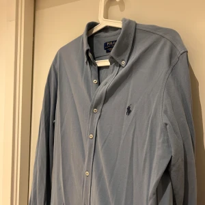 Blå skjorta från Polo Ralph Lauren - Snygg ljusblå skjorta från Polo Ralph Lauren i featherweight mesh. Klassisk design med knappar framtill och broderad logga på bröstet. Skjortan har lång ärm och en mjuk, lätt bomullskänsla som gör den riktigt skön att bära.