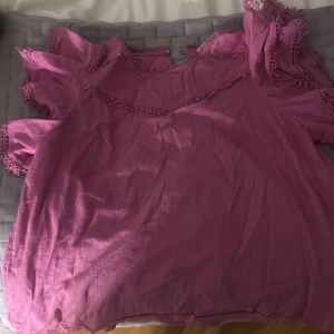 Rosa blus med volang och spetsdetaljer - Säljer en söt rosa blus med volang och spetsdetaljer vid ärmar och halsringning. Blusen har korta ärmar och en luftig passform, perfekt för dig som gillar romantiska och feminina plagg.