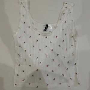 Vit blommig linnetopp från H&M Divided - Supersöt vit linnetopp från H&M Divided med små rosa blommor och diskreta ränder. Toppen har spetskant runt urringningen och en liten rosett framtill. Perfekt för en romantisk och feminin stil, och materialet är mjukt och stretchigt.