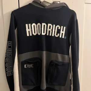 Svart och grå HOODRICH hoodie - Snygg hoodie från HOODRICH i svart och grått med stora logotryck fram, bak och på ärmen. Tröjan har två praktiska fickor med dragkedja framtill och en kontrastfärgad huva. Perfekt för dig som gillar streetwear och vill sticka ut.