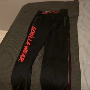 Svarta Gorilla Wear träningsbyxor L/XL - Svarta mjukisbyxor från Gorilla Wear med röd logga längs benet och på linningen. Snygga röda detaljer och dragkedjor vid fickorna. Perfekta för gymmet eller chill hemma. Skön passform och sportig look.