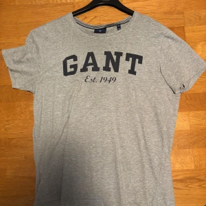 Grå GANT t-shirt med tryck - Snygg grå t-shirt från GANT i storlek M med klassiskt tryck 'GANT Est. 1949' på bröstet. Rund hals med diskreta ränder på insidan av kragen. Perfekt för en chill och stilren look, tillverkad i mjuk bomull.