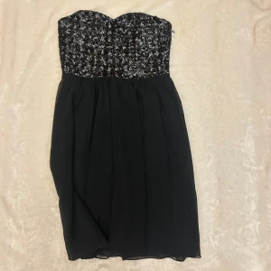 Svart paljettklänning axelbandslös - Super söt axelbandslös svart kortklänning med glittrig paljettöverdel och lätt utställd kjol i chiffongliknande tyg. Snygg sweetheart-ringning och smock baktill för extra komfort. 