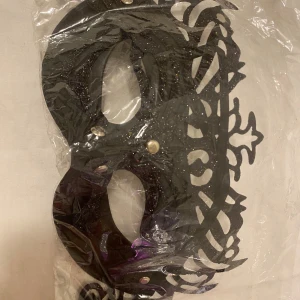 Svart glittrig mas - Snygg svart maskeradmask med glittriga detaljer och dekorativt utskuret mönster. Masken har små silvriga nitar och passar perfekt för fest eller bal. 