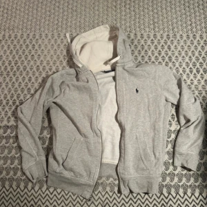Grå hoodie från Polo Ralph Lauren - Grå hoodie från Polo Ralph Lauren med dragkedja och huva med vit insida. Klassisk logga broderad på bröstet, två fickor fram och ribbade muddar. Lite sönder
