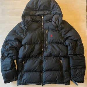 Svart pufferjacka från Polo Ralph Lauren - Svart pufferjacka från Polo Ralph Lauren med huva och flera dragkedjor. Jackan har det klassiska röda Polo-loggan broderad på bröstet och är tillverkad i syntetmaterial. Perfekt för kalla dagar med sin tjocka och quiltade design. Det saknas en knapp på luvan (syns på sista bilden), den ena stämmekanismen som man spänner midjan med är lite sönder men funkar. I övrigt är jackan i väldigt fint skick, använd i en säsong. Nypris ligger runt 6000kr.