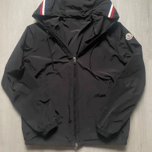 Svart vindjacka från Moncler - Svart vindjacka från Moncler med huva och dragkedja framtill. Jackan har Moncler-logga på ärmen och snygga rödvita ränder på huvan. Materialet är lätt och syntetiskt, perfekt för blåsiga dagar. Nfc och tag funkar och kommer direkt till moncler orginala hemsida. Felfri och inga skador alls. 