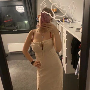 Beige långklänning med snörning - Säljer en beige långklänning med smala axelband och söt snörning framtill. Klänningen har en rak passform, slits nertill och är tillverkad i ett lätt material som känns perfekt för varmare dagar. Enkel och stilren design som är lätt att matcha.