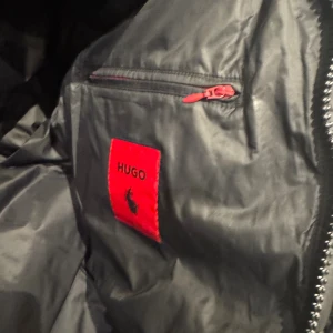 Svart pufferjacka från HUGO XXL - Svart pufferjacka från HUGO med röd logopatch på bröstet och insidan. Jackan har röda dragkedjor och är tillverkad i syntetmaterial. Perfekt för dig som vill ha en clean och modern look med extra edge. Jackan har en quiltad design och lång ärm. Jag har använt den två gånger endast kvittot finns 