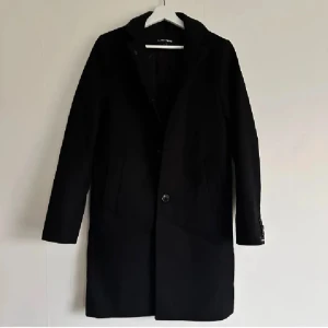 Svart rock från J.Lindeberg - Svart rock från J.Lindeberg med klassisk krage och raka linjer. Jackan har knappar framtill och fyra dekorativa knappar vid ärmslutet. Snygg och stilren design som passar perfekt till höst och vinter. Materialet är ullblandning för extra värme. Använt knappt!