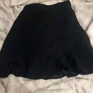 Svart kortkjol med lätt vidd - Snygg svart kortkjol med lätt utställd form. Kjolen har en enkel design utan synliga detaljer eller mönster, vilket gör den lätt att matcha med olika toppar. Perfekt för dig som gillar stilrena plagg med lite vidd nedtill.