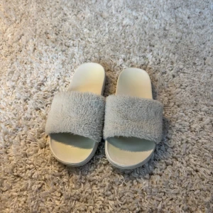 Vita fluffiga sandaler med platt sula - Säljer ett par sandaler i ljusbeige/vitt med mjukt, fluffigt band över foten. Sulan är platt och vit, med rutmönstrad undersida för extra grepp. Perfekta för chill dagar hemma eller när du vill ha något bekvämt och mysigt på fötterna. strlk 38/39