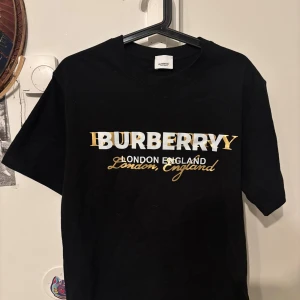 Svart Burberry t-shirt med tryck - Svart t-shirt från Burberry med stort vitt och guldigt tryck på bröstet där det står 'BURBERRY London England'. Klassisk rund hals och korta ärmar. Materialet är mjuk bomull och passformen är normal. Perfekt för dig som gillar stilrena designer med lyxig känsla.
