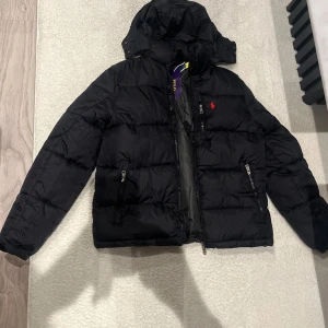 Svart pufferjacka från Polo Ralph Lauren - Svart pufferjacka från Polo Ralph Lauren med röd broderad logga på bröstet. Jackan har huva, två sidofickor med dragkedja och quiltad design. Perfekt för kalla dagar och har en clean, modern look.