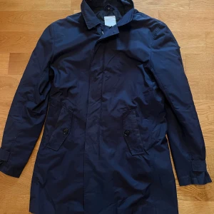 Marinblå rock från J.Lindeberg - Snygg marinblå rock från J.Lindeberg med klassisk krage och två framfickor med knappar. Jackan har en stilren design med metallmärke på kragen och diskret dragkedja under knappslå. Perfekt för dig som gillar cleana och tidlösa plagg.