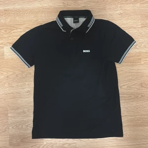 Svart slim fit t-shirt från BOSS - Säljer en svart t-shirt från BOSS med storlek S. Slim fit stretch-modell med korta ärmar och vit-randiga detaljer på krage och ärmslut. Hör av er vid intresse! 