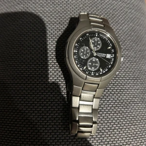 Citizen Titanium herrklocka svart urtavla - Stilren Citizen herrklocka i titan med svart urtavla och flera små urtavlor för extra funktioner. Klockan har datumvisning och robust länkarmband i mattgrått titan. Perfekt för dig som gillar en sportig och klassisk look med tekniska detaljer.