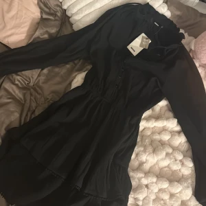 Svart volangklänning från Bik Bok - Supersnygg svart kortklänning från Bik Bok med volanger och knappar framtill. Klänningen har långärmade puffärmar, markerad midja och ett skirt, lätt material som ger en flowig känsla. 