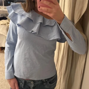 Ljusblå blus med volang - Supersöt ljusblå blus med stora volanger över axlarna och bröstet. Blusen har lång ärm med knapp vid ärmslut och är tillverkad i ett mjukt bomullstyg. Perfekt för dig som gillar romantiska detaljer och en feminin look.