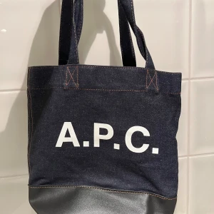 Mörkblå shoppingväska från A.P.C. - Snygg mörkblå shoppingväska från A.P.C. i denim med svart botten i skinn. Väskan har rejäla handtag, vit logotyp på framsidan och en innerficka med dragkedja. Perfekt storlek för att få med allt du behöver och stilren design.