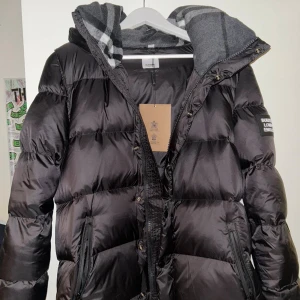 Svart pufferjacka från Burberry - Svart pufferjacka från Burberry med huva och klassiskt rutigt foder. Jackan har dragkedja och knappar framtill, samt två sidofickor med dragkedja. Perfekt för kalla dagar med sin fluffiga och isolerande design.