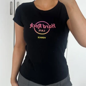 Topp 🖤 - Svart topp från Hard Rock Cafe Venice med tryck i rosa och gult framtill. Modellen är figurnära med rund hals och korta ärmar. Tillverkad i 100% bomull. Storlek L, tror det är barnstorlek, jag har XS/S och den passar mig 🖤