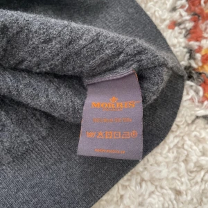 Grå kabelstickad tröja Morris xs eftersom den har krympt - Snygg mörkgrå kabelstickad tröja från Morris i 100% merinoull. Rund halsringning med röd insida och diskret röd logga broderad på bröstet. Perfekt för dig som gillar klassisk stil med en twist. Långärmad och mjuk känsla.