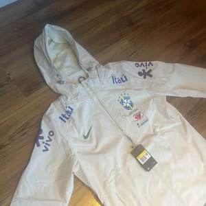  Brasil Nike vindjacka S - Säljer en vit/biege Brasil Nike vindjacka med huva och dragkedja. Jackan har Brasil-logga, grönt Nike-märke och flera tryck i blått och lila på bröst och ärmar. Materialet är lätt polyester och jackan har meshfoder. Perfekt för dig som gillar sportig stil. För mer bilder eller frågor så är  de bara att skriva👌🏼färgen är inte helt vit utan lite biege som ni ser på bilden när jag har vit t shirt under!