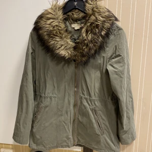 Olivgrön parkajacka med fuskpäls L.O.G.G - Parkajacka från L.O.G.G i olivgrönt med stor fluffig fuskpäls runt kragen. Jackan har dragkedja framtill, två fickor och långärmade ärmar. Perfekt för dig som gillar chill och snygg streetstil med lite extra edge. Storlek 34 men jag tycker att den är stor i storleken. Pälskragen går att knäppa bort om man inte vill ha den på 