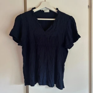 Mörkblå blus med v-ringning - Säljer en mörkblå blus med korta ärmar och v-ringad hals. Blusen har en lätt skrynklig struktur och mjukt fall. Perfekt för dig som gillar en enkel men stilren look. Storlek L men krympt i tvätten så mer en s