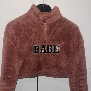 Fluffig croptop  - Supermjuk och fluffig rosa fleece-tröja med croppad passform och halv dragkedja framtill. Stort broderat BABE-tryck i svart och vitt på bröstet. Perfekt för dig som gillar cozy vibes och statement-plagg.Super mjukt och 