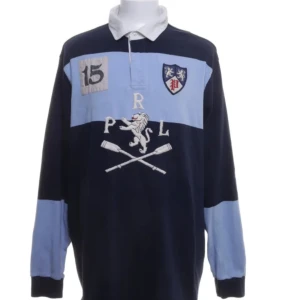 Blå rugbytröja  Polo Ralph Lauren XXL - Snygg rugbytröja från Polo Ralph Lauren i marinblått och ljusblått med broderade detaljer, lejon och åror på bröstet samt patch med siffran 15. Klassisk krage och långärmad modell, perfekt för dig som gillar sportig och preppy stil.