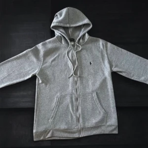 Grå hoodie från Polo Ralph Lauren - Snygg grå hoodie från Polo Ralph Lauren med klassisk broderad logga på bröstet. Tröjan har huva med snörning, dragkedja framtill och två praktiska fickor. Mjuk bomullsmix som är perfekt för chill dagar. Passar dig som gillar stilren och enkel design.