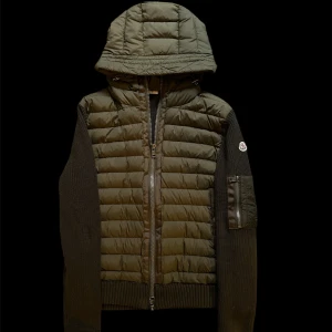 Moncler cardigan  - Snygg grön pufferjacka från Moncler med stickade ärmar och quiltad framsida. Jackan har en stor huva, dragkedja framtill och en ficka med dragkedja på ärmen. Moncler-logga på vänster ärm. Perfekt för dig som gillar street och exklusiv stil.