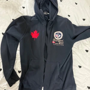 Svart hoodie Geographical Norway - Svart hoodie från Geographical Norway med dragkedja, huva och broderade detaljer. Stor röd lönnlövspatch på bröstet och broderad expedition-logga. Praktiska fickor framtill och mjukt material, perfekt för kyliga dagar.