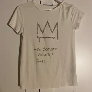 Vit t-shirt HOV1 - Vit t-shirt med glittrig krona och texten 'ni dansar vidare i livet' framtill. Klassisk rund halsringning och korta ärmar. Perfekt för dig som gillar stilrena plagg med en twist. Har själv satt på diamanterna med diamanttejp, så det går enkelt att plocka av