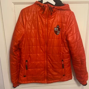 Röd quiltad pufferjacka med huva - Snygg röd pufferjacka med quiltad design och huva. Jackan har dragkedja framtill, två fickor med dragkedja och ett coolt emblem på bröstet. Perfekt för dig som vill sticka ut med färg och stil under kyliga dagar.