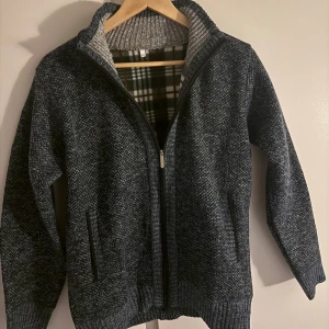 Mörkgrå stickad cardigan jacka - Mysig mörkgrå stickad cardigan jacka med dragkedja och ribbade muddar. Insidan har ett rutigt foder i svart, vitt och brunt som ger extra stilpoäng. Perfekt för kyliga dagar.