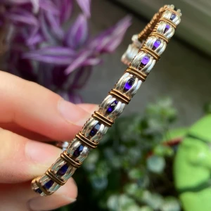 Unikt silver- och bronsfärgat armband - Snyggt armband med silver- och bronsfärgade detaljer och lila stenar infattade i varje länk. Designen är stilren med ett coolt rutmönster och metalltråd som ger en modern vibe. Perfekt för dig som gillar att sticka ut med accessoarer som har lite edge.