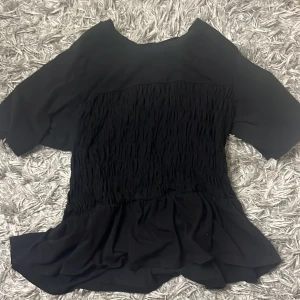 Svart blus med volang från Vero Moda - Svart blus från Vero Moda i storlek XL. Tröjan har krympt i tvätten så personligen tycker jag att den har blivit som storlek M. Toppen har korta ärmar, ett snyggt rynkat parti över bröstet och en bred volang .